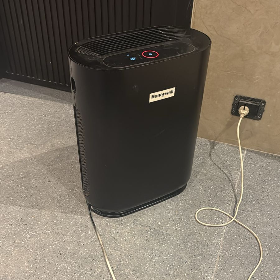 Black air purifier on a concrete floor with a power cord plugged into an outlet.